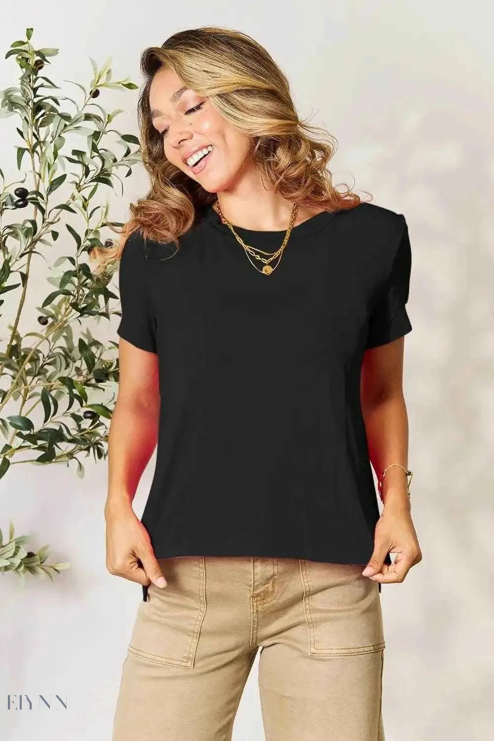 Smiling model wearing a women's basic black t-shirt with crew neck and short sleeves paired with tan pants