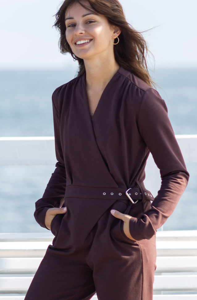 Woman wearing a brown outfit with a belt, standing against a blurred outdoor background.